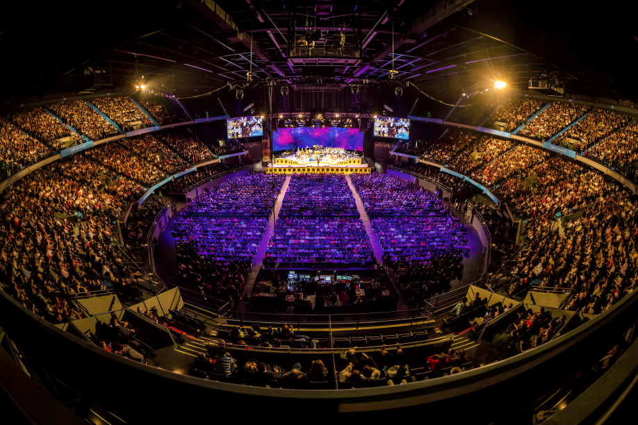 Vista interior del Movistar Arena en Bogotá lleno de gente durante un concierto, con luces de escenario y pantallas gigantes.
