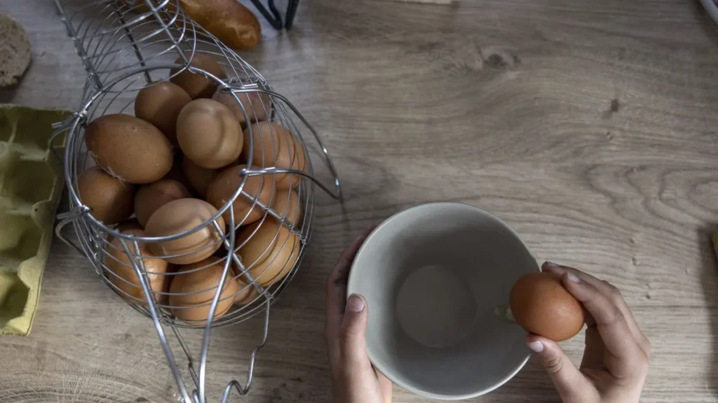 Mujer rompiendo un huevo fresco en un bol aparte, con una caja de huevos y utensilios de cocina limpios en el fondo.