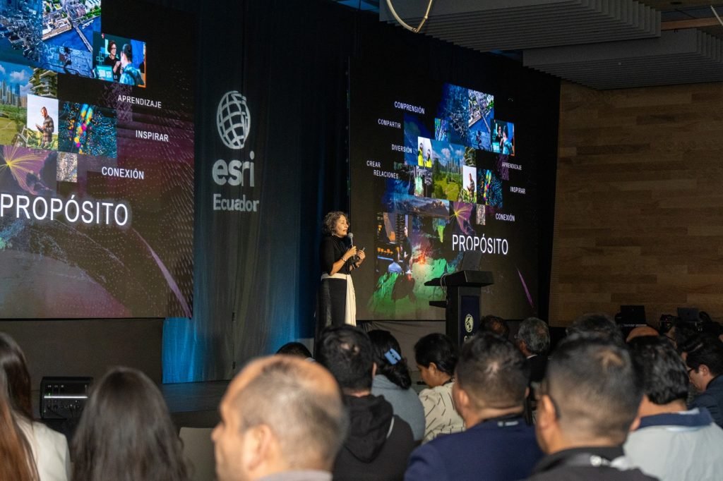 Conferencia de Esri Ecuador mostrando una pantalla con un mapa geoespacial interactivo y personas asistiendo al evento, destacando la infraestructura geoespacial y la inteligencia artificial.