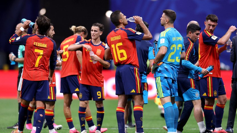 Jugadores de fútbol reunidos durante una pausa para hidratación en el Mundial, con árbitros y personal técnico cerca, en un estadio con espectadores al fondo