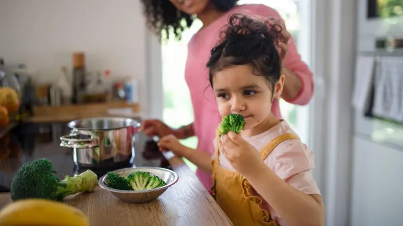 Niña pequeña oliendo un trozo de brócoli en la cocina, ilustración sobre estrategias para que los niños coman verduras
