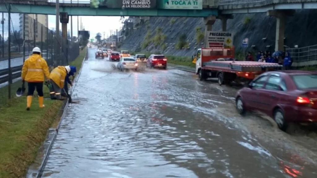 Trabajadores con impermeables limpian una vía anegada mientras circulan vehículos bajo la lluvia